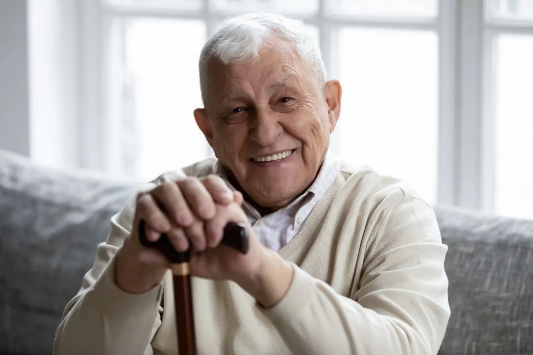 Elderly man with white hair smiling while holding a walking cane, seated on couch indoors.