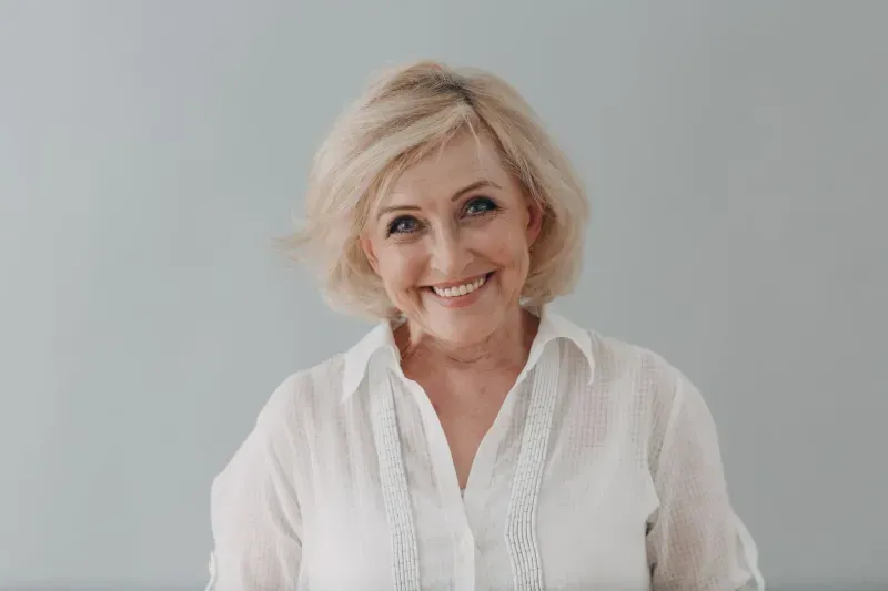 Professional headshot of mature woman with blonde hair wearing white blouse against neutral background.