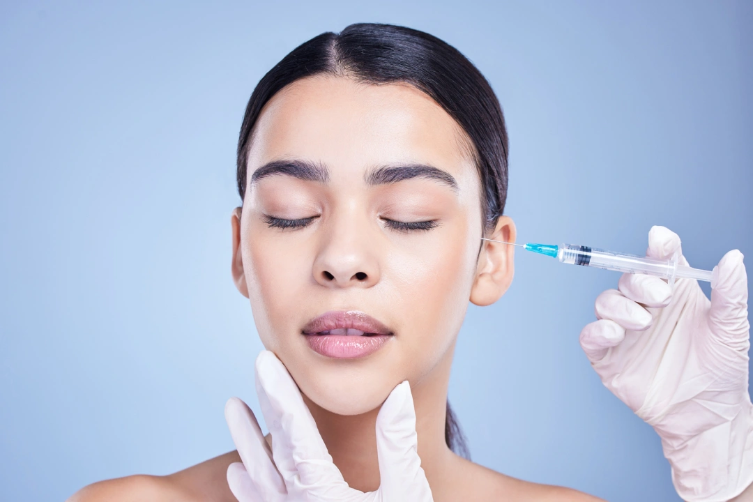 Woman receiving cosmetic injection on cheek against blue background in medical setting.