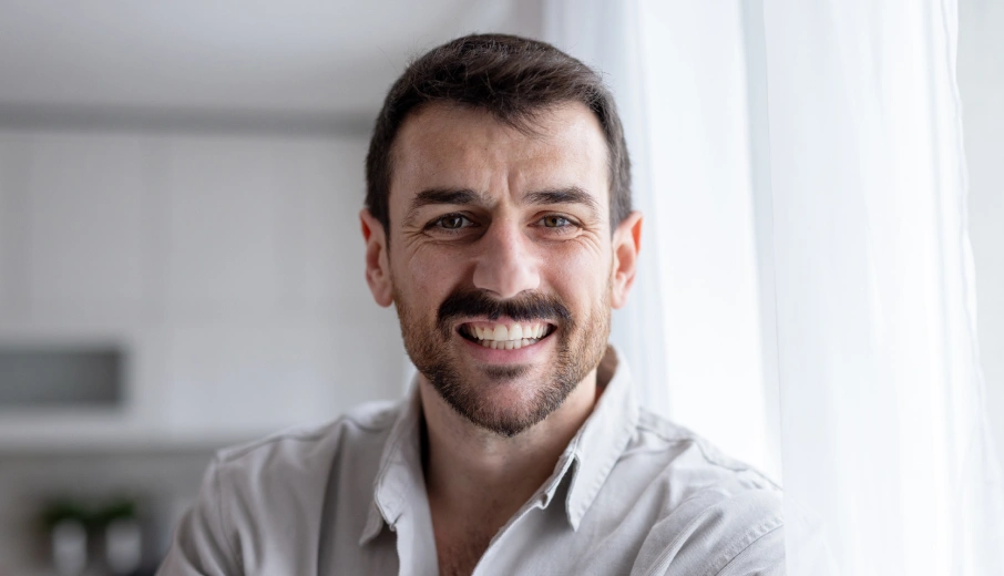 Smiling male dentist in light blue shirt, professional headshot in modern clinic setting.