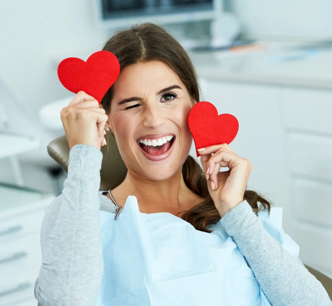 Woman holding red hearts in front of face, smiling in bright dental office setting.