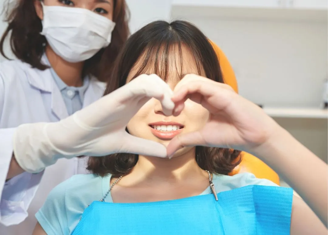 Young patient making heart shape with hands while wearing blue bib in dental office with masked dentist behind her.